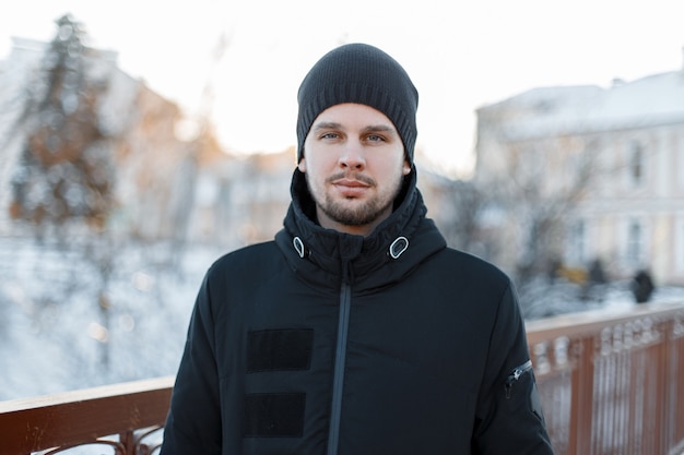 Retrato de un elegante joven atractivo con barba en una elegante chaqueta negra de invierno con un gorro de punto en un cálido día de invierno. Chico brutal de moda.