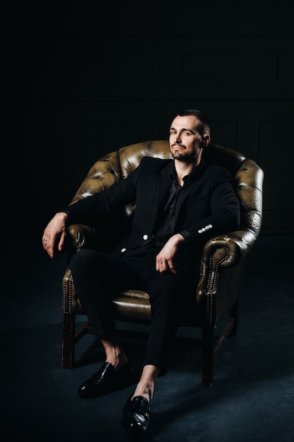 Retrato de un elegante joven europeo con un traje sentado en una silla en un interior oscuro.