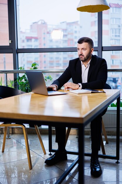 Retrato de un elegante joven hombre de negocios, en una gran oficina moderna en lo alto de un piso superior, mirando a una computadora portátil. Hombre de negocios en el escritorio de la oficina de finanzas freelance