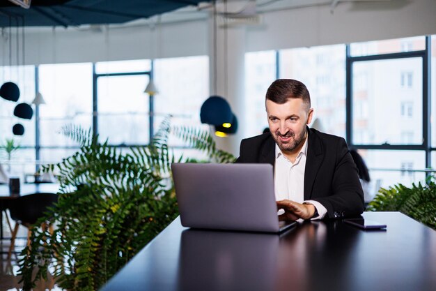 Retrato de un elegante joven hombre de negocios, en una gran oficina moderna en lo alto de un piso superior, mirando a una computadora portátil. Hombre de negocios en el escritorio de la oficina de finanzas freelance