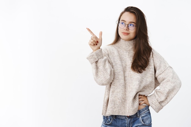 Retrato de elegante joven mandona con gafas y suéter apuntando a la esquina superior izquierda y mirando con expresión creativa inteligente