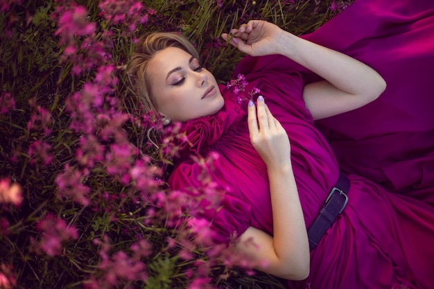 Retrato elegante linda jovem deitada em um vestido rosa em um campo com flores silvestres no verão ao pôr do sol