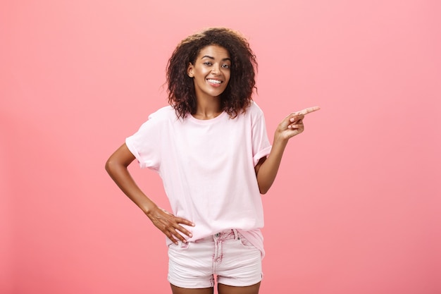 Retrato de una elegante mujer afroamericana con peinado afro sosteniendo la mano en la cintura apuntando a la derecha y sonriendo con alegría.