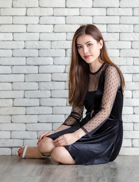 Retrato de elegante mujer asiática, etnia lao, en elegante vestido negro sentado en el suelo y mirando a la cámara con una sonrisa brillante y pose positiva sobre fondo de pared de ladrillo