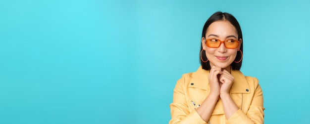 Retrato de una elegante mujer coreana con gafas de sol sonriendo y mirando a un lado el logo coqueto de pie sobre fondo azul Copiar espacio