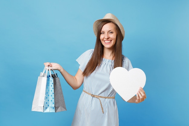 Retrato elegante mulher bonita com vestido de verão, chapéu de palha segurando pacotes sacos com compras depois de fazer compras, coração branco com espaço de cópia para anúncio isolado em fundo azul pastel.