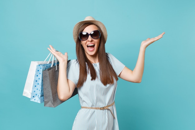 Retrato elegante mulher feliz atraente com vestido de verão, chapéu de palha, óculos de sol segurando sacolas de pacotes com compras depois de fazer compras isoladas em fundo azul pastel. copie o espaço para anúncio.