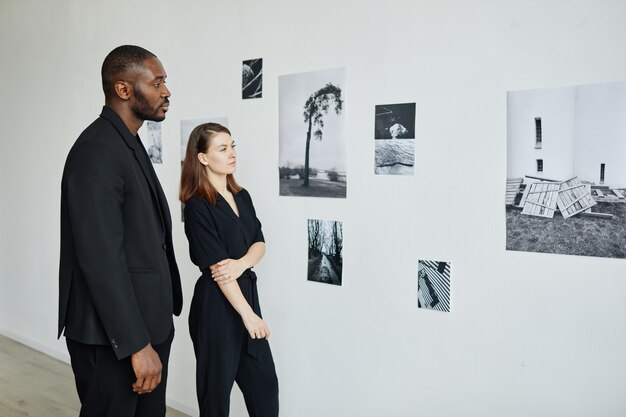 Retrato de una elegante pareja de raza mixta vestida de negro mientras mira fotografías en la galería de arte moderno, espacio de copia