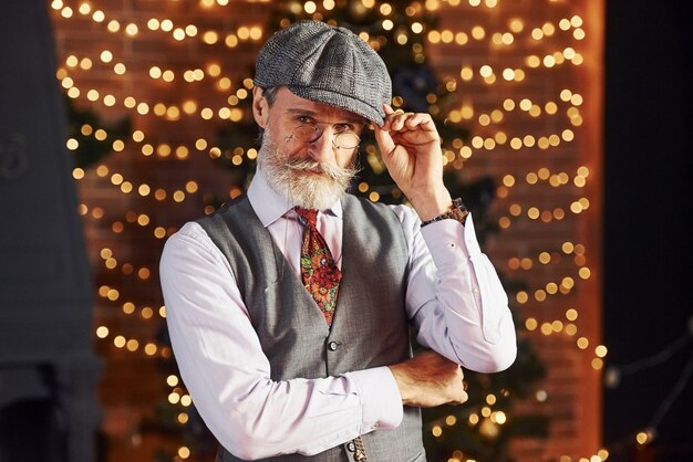 Retrato de elegante senior con cabello gris y barba en chaqueta y camisa blanca está en la sala de Navidad decorada.