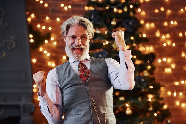 Foto retrato de elegante senior con cabello gris y barba en chaqueta y camisa blanca está en la sala de navidad decorada.