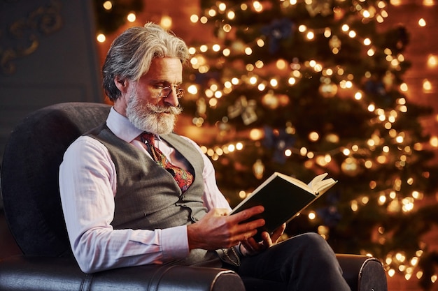 Retrato de elegante senior con cabello gris y barba libro de lectura en la sala de Navidad decorada.