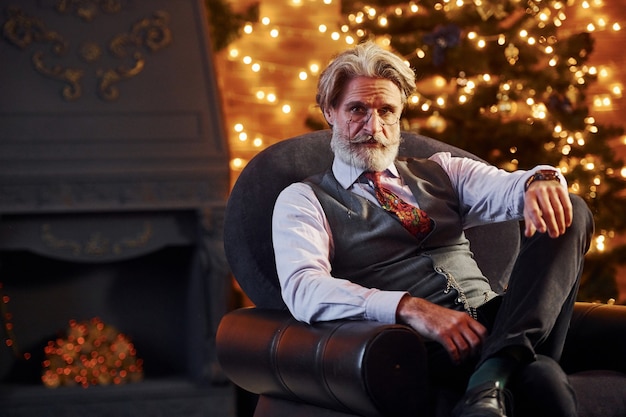 Retrato de elegante senior con canas y barba está en la sala de Navidad decorada sentado en la silla.