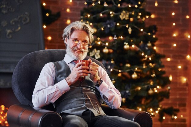 Retrato de elegante senior con canas y barba está en una sala de Navidad decorada con una taza de bebida.