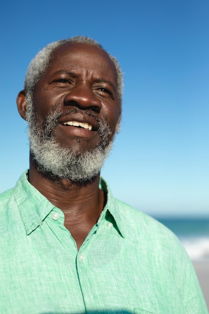 Retrato em close de um homem afro-americano sênior de pé na praia com o céu azul e o mar ao fundo, olhando para longe