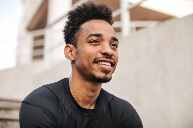 Retrato em close-up de um homem moreno encaracolado de pele escura em uma camiseta de manga comprida sorrindo sinceramente e posando ao ar livre