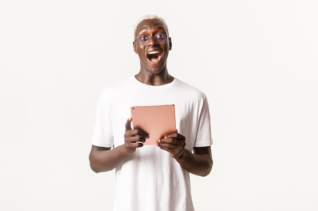 Retrato de emocionado apuesto estudiante afroamericano en gafas, mirando asombrado, sosteniendo tableta digital