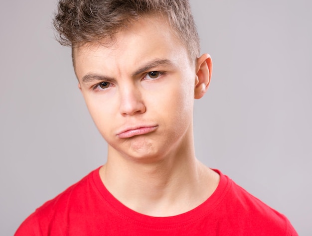 Retrato emocional de um adolescente caucasiano com problemas tristes, um menino triste olhando para a câmera, uma criança preocupada vestindo uma camiseta vermelha em fundo cinza.