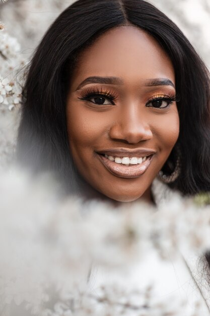 Retrato emocional feliz de uma bela jovem negra com um sorriso em flores na natureza em um dia de primavera. Linda garota africana sorri e gosta de árvores floridas