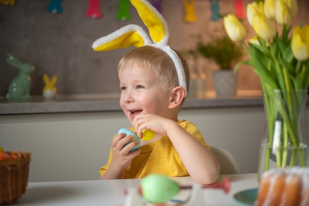 Retrato emocional de un niño alegre con orejas de conejo el día de Pascua que se ríe alegremente juega con coloridos huevos de Pascua sentados en una mesa en la cocina