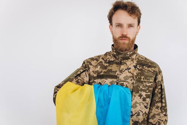 Retrato de un emotivo joven soldado patriota ucraniano barbudo con uniforme militar sosteniendo una bandera en la oficina