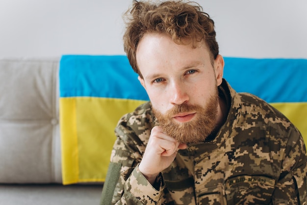 Retrato de un emotivo joven soldado patriota ucraniano con uniforme militar sentado en la oficina en un sofá con una bandera amarilla y azul