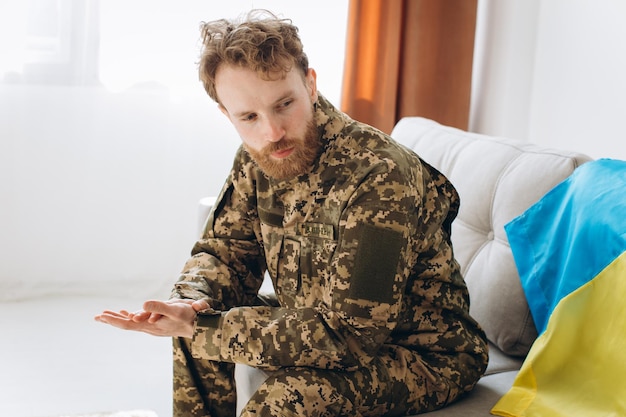 Retrato de un emotivo joven soldado patriota ucraniano con uniforme militar sentado en la oficina en un sofá con una bandera amarilla y azul