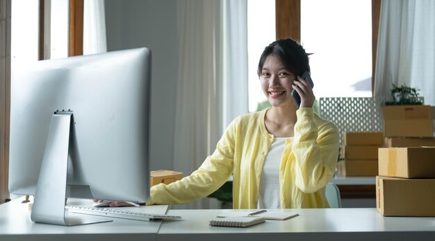 Un retrato de una empleada asiática de comercio electrónico sentada en la oficina llena de paquetes en la mesa usando una computadora portátil y un teléfono inteligente para la tecnología de comercio electrónico de PYME y el negocio de entrega