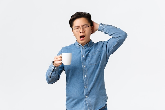 Retrato de empleado masculino joven soñoliento bostezando y sosteniendo la taza con café, despertando cansado. Hombre trabajando hasta tarde con ganas de dormir, sosteniendo la cabeza y los ojos cerrados, de pie fondo blanco.