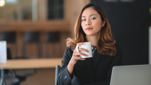 Retrato de empresaria asiática trabajando en su proyecto y tomando un café mientras mira a la cámara