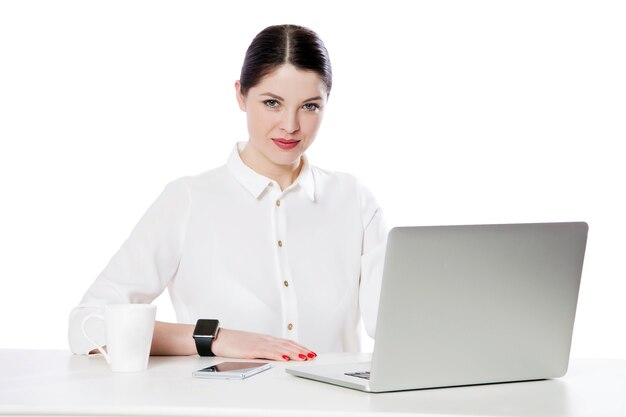 Retrato de empresaria morena atractiva tranquila exitosa con maquillaje en camisa blanca sentado con portátil y mirando a cámara con una sonrisa. tiro del estudio de interior, aislado en fondo blanco.