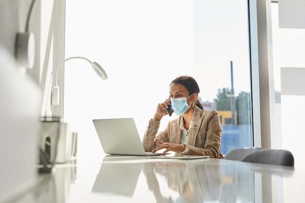 Retrato de empresaria de raza mixta con máscara y hablando por teléfono inteligente mientras trabaja en la oficina blanca, espacio de copia