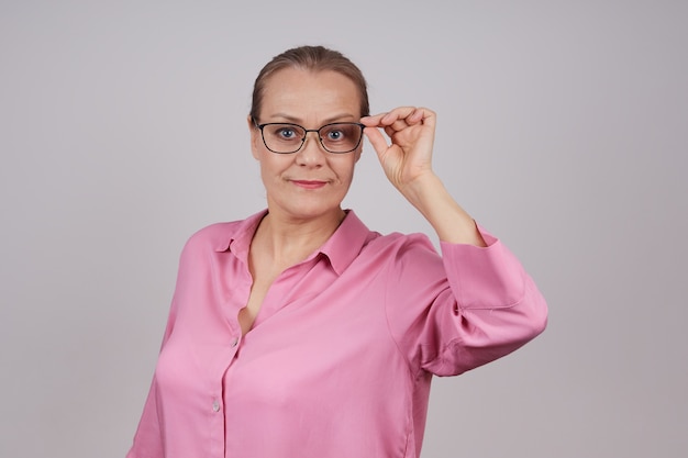 Retrato de una empresaria senior en una blusa rosa sosteniendo el arco de sus gafas con la mano. Foto aislada sobre un fondo gris con espacio de copia.