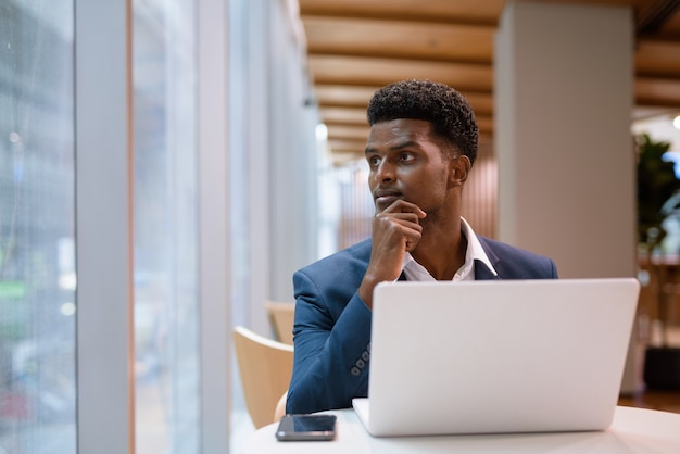 Retrato del empresario africano con ordenador portátil en la cafetería mientras mira por la ventana, disparo horizontal