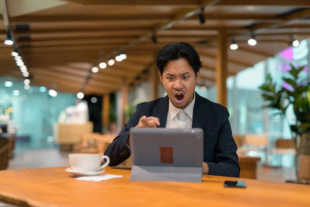 Retrato del empresario asiático enojado en la cafetería con tableta digital