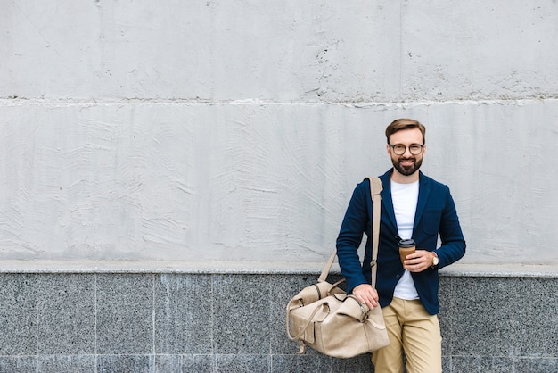 Retrato del empresario barbudo con anteojos sosteniendo un vaso de papel y una bolsa de transporte mientras está de pie cerca de la pared