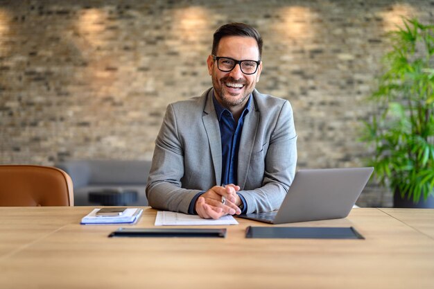 Retrato de un empresario guapo y sonriente con una computadora portátil y documentos en el escritorio sentado en una oficina moderna