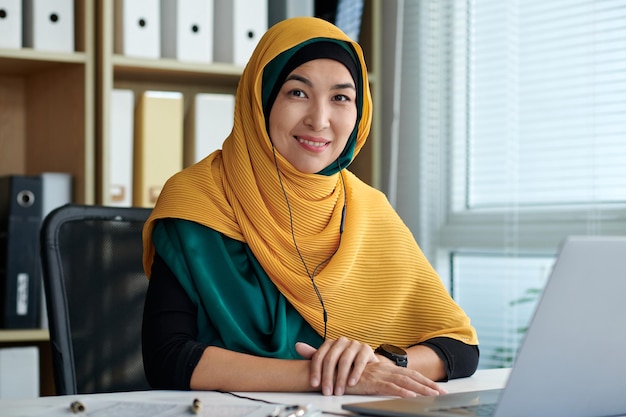 Retrato de un empresario musulmán sonriente con hijab amarillo sentado en el escritorio de la oficina