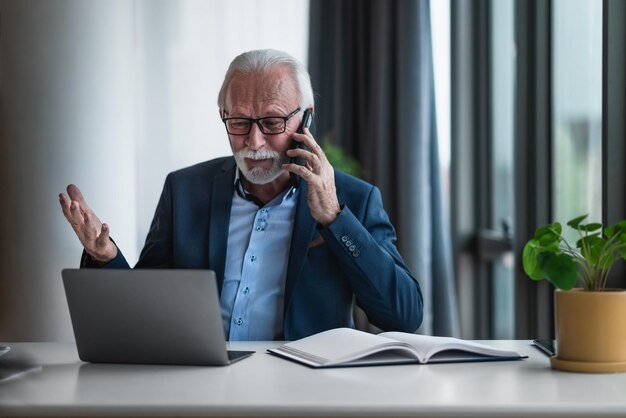 Retrato de un empresario senior maduro en una oficina de negocios informal que hace llamadas telefónicas y gesticula mientras trabaja con una computadora portátil y examina la búsqueda de datos e informes