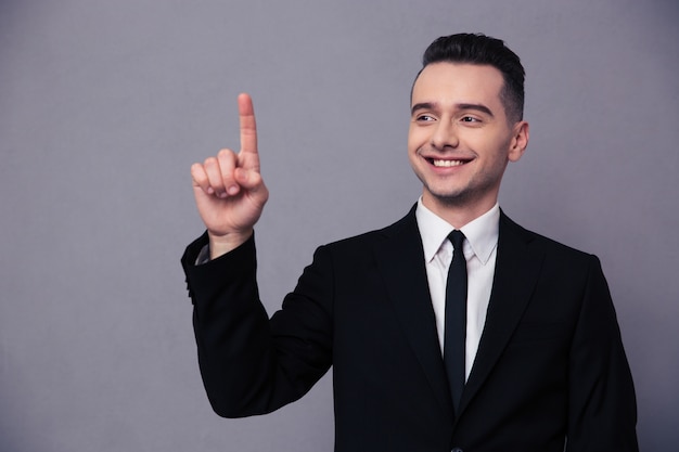 Retrato de un empresario sonriente apuntando con el dedo hacia arriba sobre la pared gris