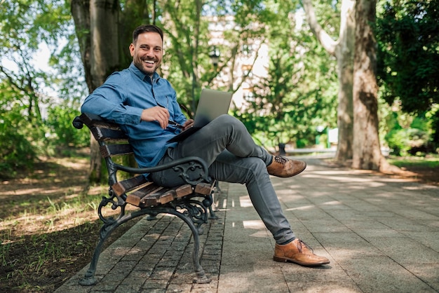 Retrato de un empresario sonriente que usa una laptop mientras se sienta en un banco en el parque