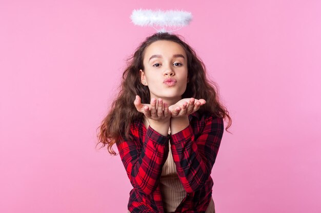 Retrato de una encantadora adolescente con cabello rizado de morena en camisa a cuadros y halo de juguete sobre la cabeza enviando un beso de aire con labios fruncidos, romance en el día de san valentín. tiro del estudio aislado sobre fondo de color rosa