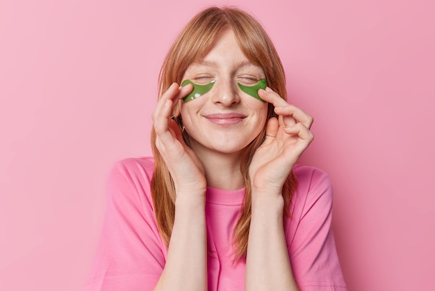 Retrato de una encantadora chica pelirroja pecosa que se pone parches de hidrogel verde debajo de los ojos para el tratamiento de la piel viste una camiseta informal aislada sobre fondo rosa Concepto de belleza, bienestar y cuidado facial