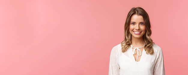 Retrato de una encantadora chica rubia con un hermoso vestido blanco con una sonrisa radiante mirando a la cámara sintiéndose feliz y alegre disfrutando de los soleados días románticos de primavera con un fondo rosa