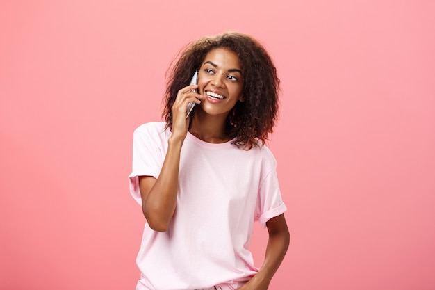 Retrato de encantadora joven afroamericana con peinado afro sosteniendo smartphone y mirando a la izquierda con curiosidad.