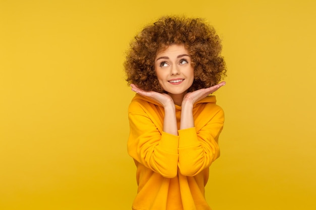 Retrato de una encantadora joven juguetona de cabello rizado con capucha de estilo urbano que muestra su rostro limpio y fresco con el concepto de belleza maquillada y cuidado de la piel en un estudio interior aislado en un fondo amarillo