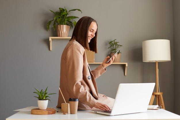 Retrato de una encantadora mujer de cabello oscuro feliz y satisfecha con una chaqueta beige posando en la oficina trabajando en una laptop y sosteniendo un teléfono móvil disfrutando de su trabajo sentada en la mesa