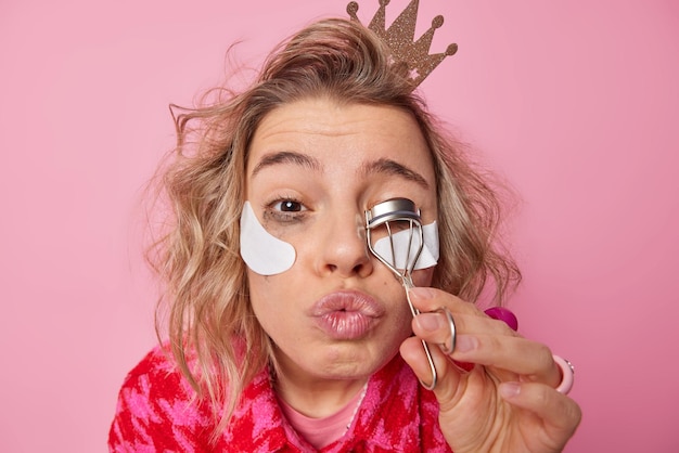 Retrato de una encantadora mujer europea que se maquilla usa rizador de pestañas mantiene los labios doblados aplica parches de belleza debajo de los ojos tiene peinado lleva una pequeña corona en la cabeza aislada sobre fondo rosa