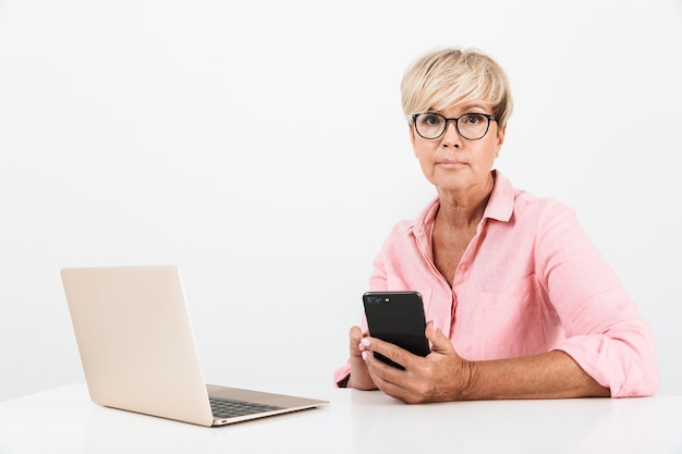 Retrato de una encantadora mujer de mediana edad con anteojos sosteniendo un teléfono celular y usando una computadora portátil aislada sobre una pared blanca