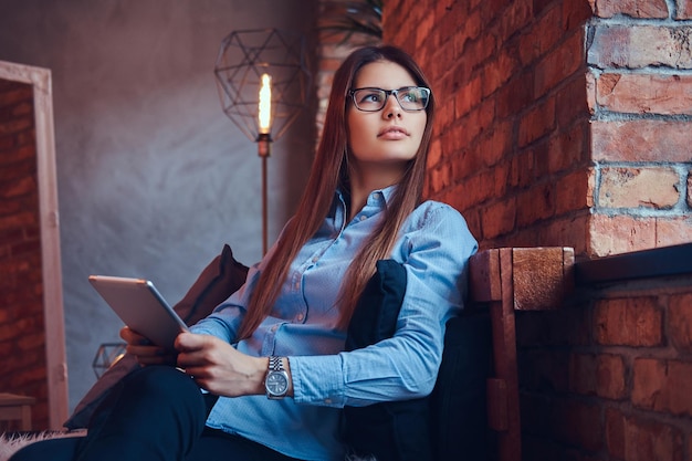 Retrato de una encantadora mujer de negocios morena con gafas y camisa azul sostiene una tableta y se sienta en un sofá en una habitación con diseño de loft.