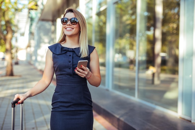 Retrato de una encantadora mujer de negocios parada con una maleta y usando un teléfono móvil